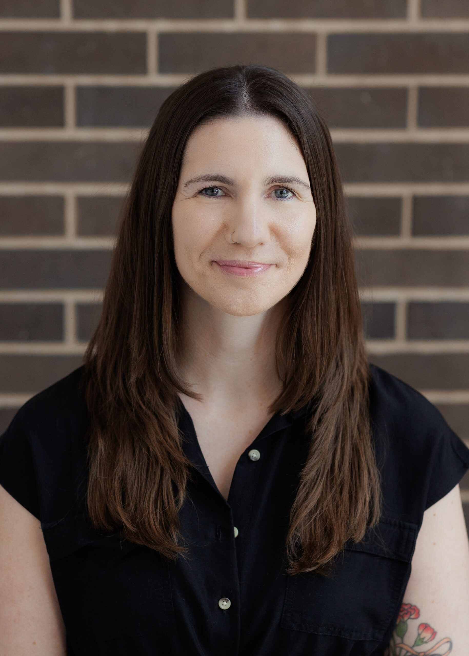 A woman with long brown hair wearing a black collared shirt posing in front of a brick wall.