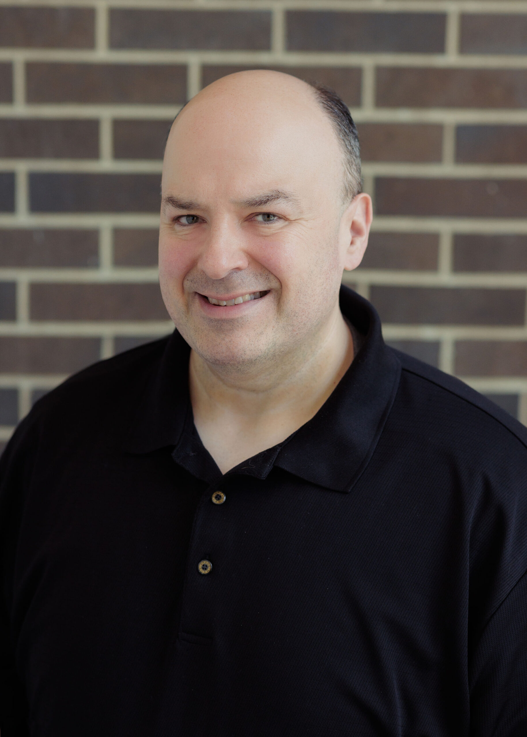 A man with a shaved head smiles while wearing a black polo shirt in front of a brick wall.