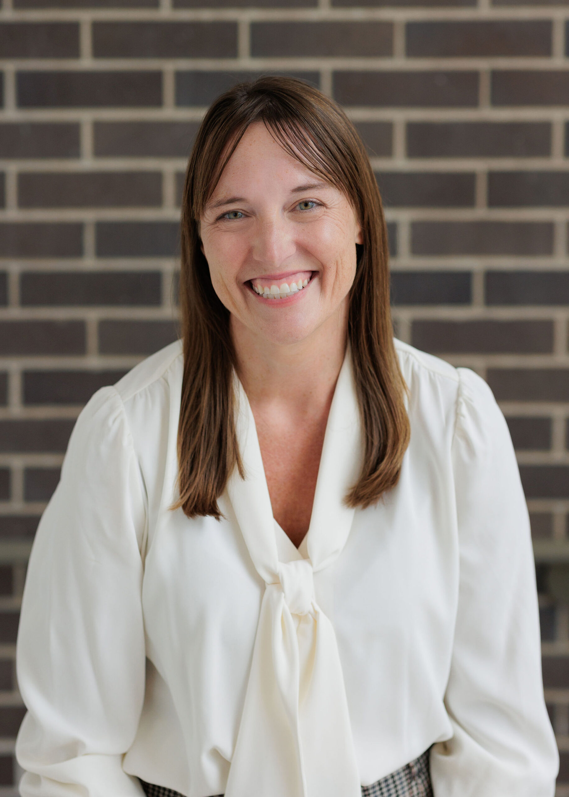 A smiling woman with brown hair wearing a white blouse against a dark brick wall.