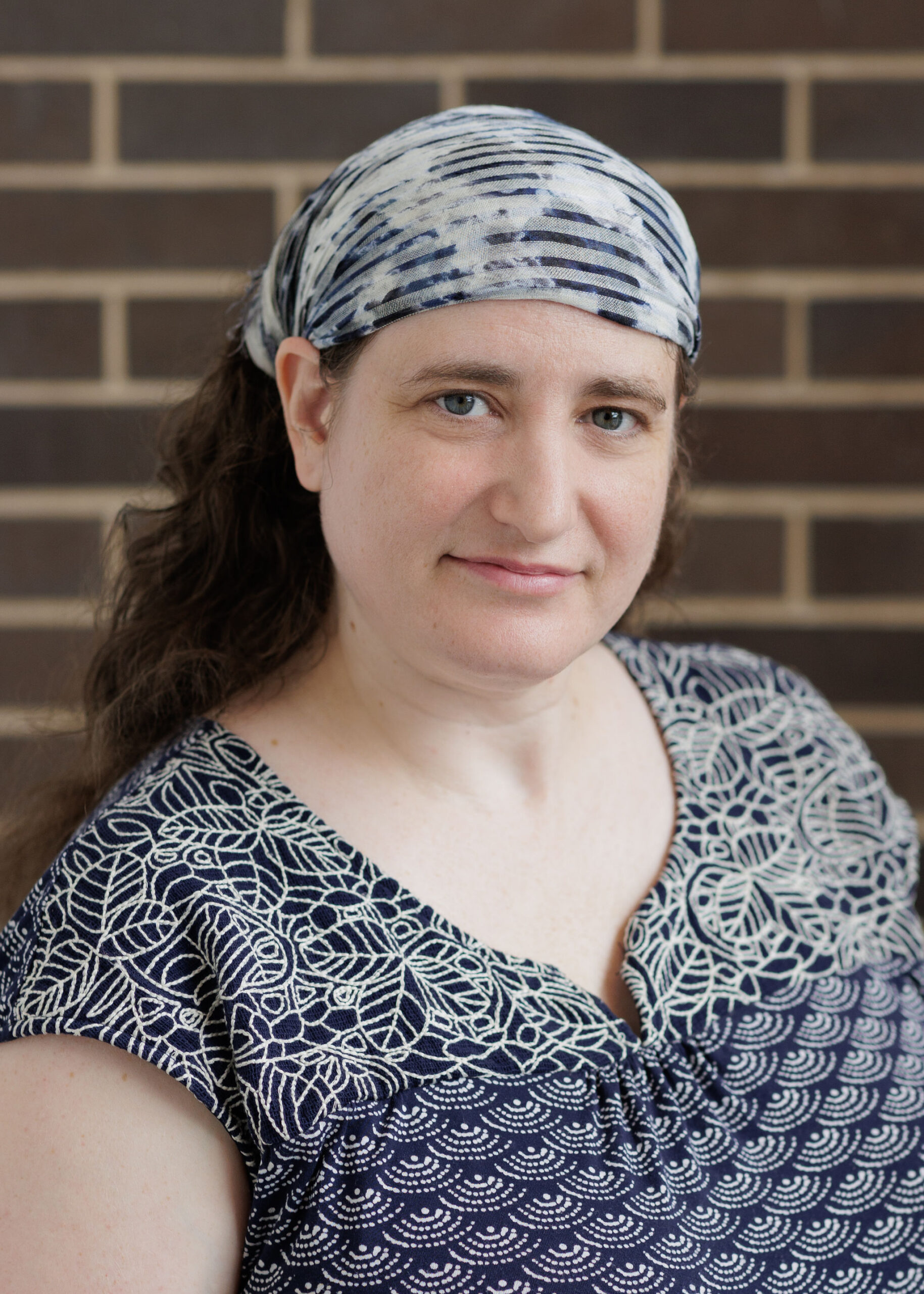 A woman wearing a patterned headscarf and a navy blue patterned top poses in front of a brick wall.