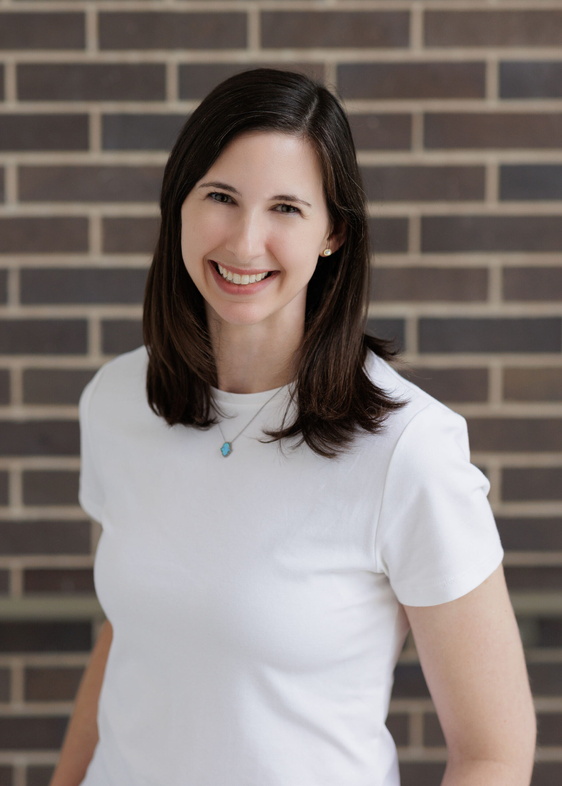 A woman with shoulder-length dark hair wearing a white t-shirt and a small blue pendant necklace smiles at the camera in front of a brick wall.