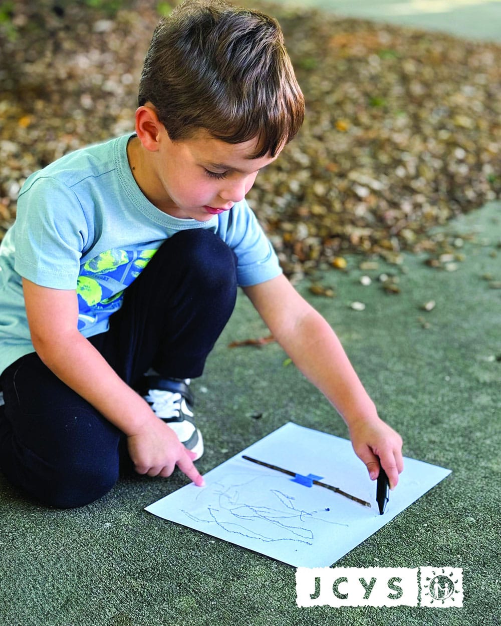 A young boy kneels on outdoor pavement, pointing to a drawing on a white sheet of paper with one hand and holding a black marker in the other. The JCYS logo appears in the lower right corner of the image.
