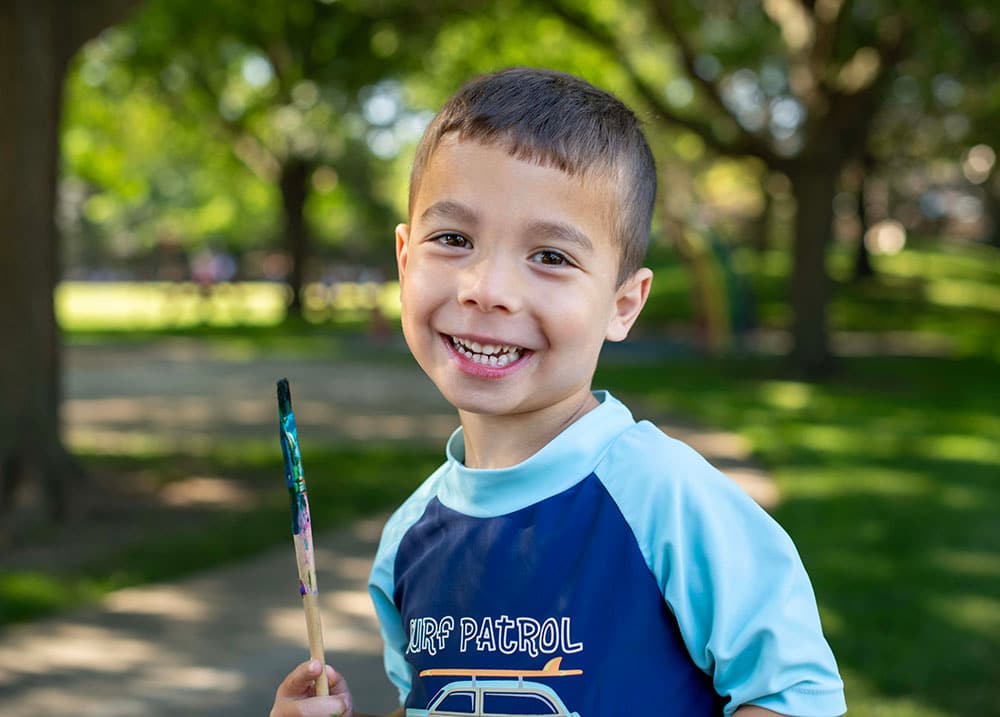 Smiling young boy holding a paintbrush stands outside in a sunny park. He is wearing a blue Surf Patrol shirt. Green grass and trees are visible in the blurred background.