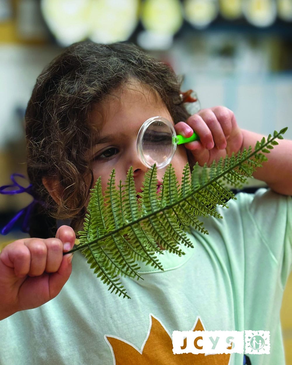A young child holds a green fern and looks at it through a magnifying glass. The child wears a light-colored shirt, and the background is blurred. The JCYS logo appears in the lower right corner.