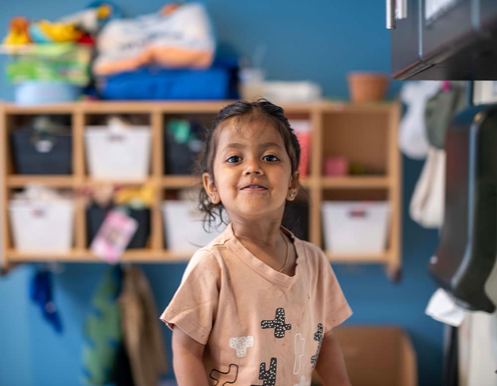 A young child with long hair stands indoors, wearing a light pink shirt with graphic designs. Behind them are shelves with bins and various items against a blue wall.