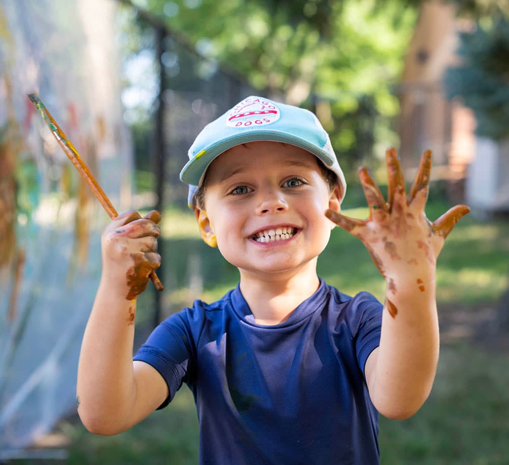 A young child wearing a blue shirt and light blue cap smiles while holding a paintbrush in one hand and showing their paint-covered hands outdoors, with blurred greenery and sunlight in the background.