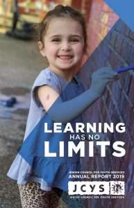 A young girl smiles while painting a mural on a wall. She holds a small brush, wearing a light purple shirt and leopard print pants. The text on the image reads, Learning has no limits. JCYS Annual Report 2019..