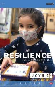 Young child wearing a patterned face mask and navy shirt, holding a book. Text on image reads, RESILIENCE JCYS ANNUAL REPORT | 2021. JCYS logo and QR code are also visible at the bottom.