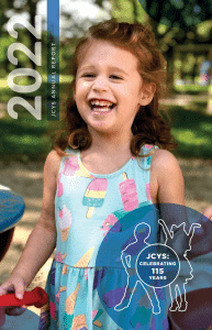 Young child smiling outdoors, wearing a sleeveless dress with colorful ice cream prints. The image includes 2022 JCYS Annual Report text and a blue graphic stating JCYS: Celebrating 115 Years. Trees and playground equipment in background.