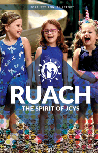 Three young children wearing colorful dresses stand outside, smiling and laughing. The cover text reads 2023 JCYS Annual Report with RUACH: The Spirit of JCYS overlaid on the image, along with a circular logo.