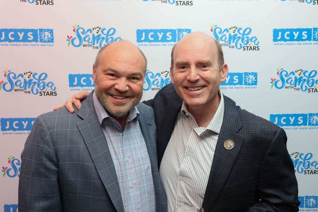 Two men in suits smile in front of a step-and-repeat banner with logos, including JCYS and Samba with the Stars. One man has his arm around the others shoulder. Indoor event setting.