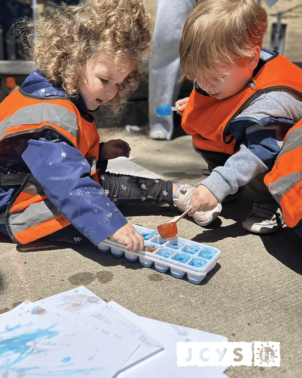 Two young children wearing orange safety vests sit on the ground, using popsicle sticks to mix blue liquid in an ice cube tray. Papers with blue marks and the JCYS logo are visible nearby.