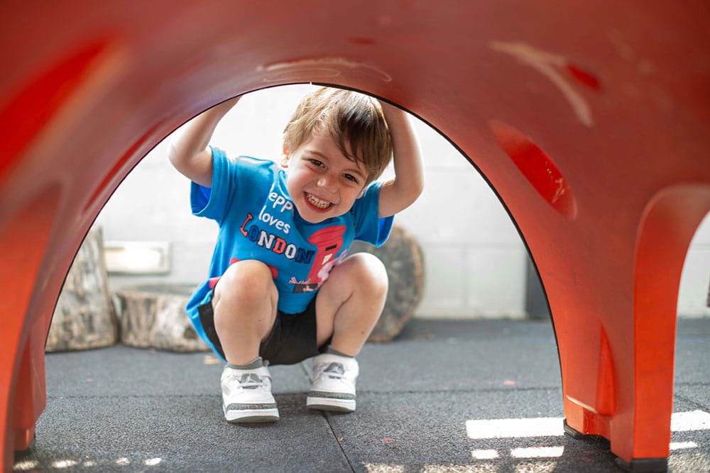 A young child crouches and smiles under a large red playground structure, holding onto the sides. The child wears a blue shirt, black shorts, and white sneakers. The background includes tree stumps and a light wall.