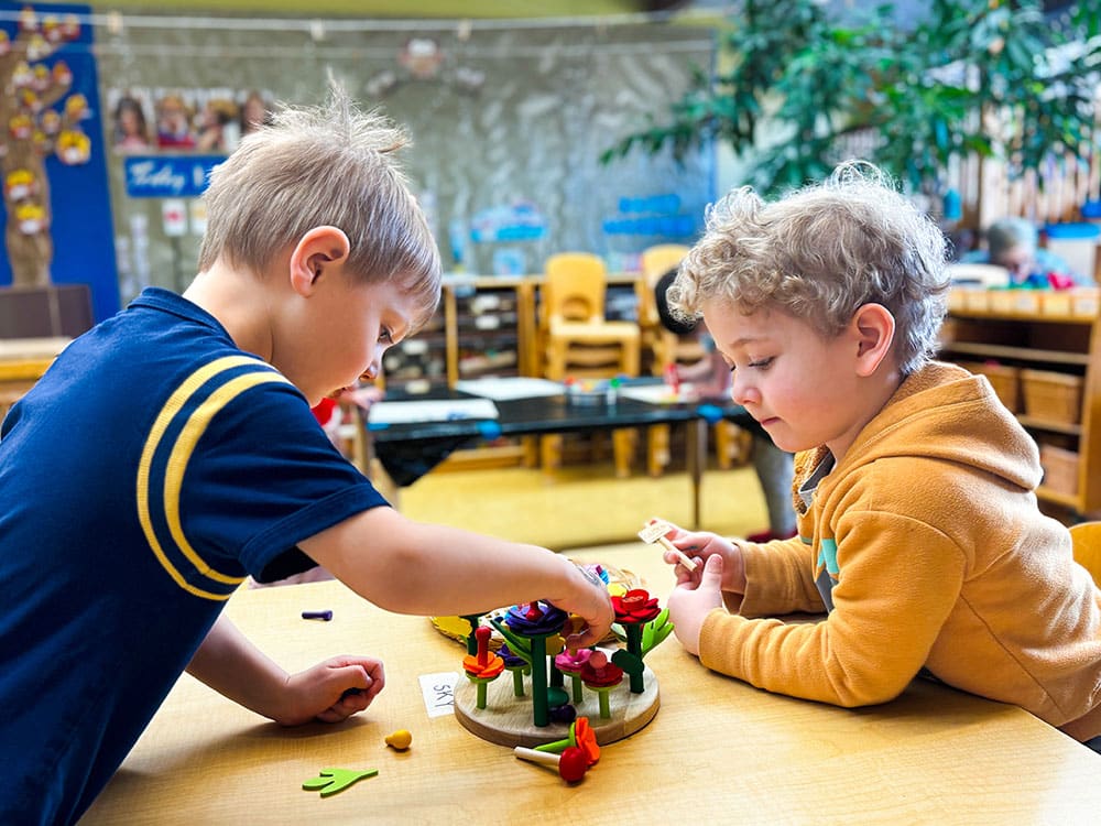 Two young children sit at a table in a classroom, playing with a colorful flower-building toy. The background shows chairs, shelves, and classroom decorations.