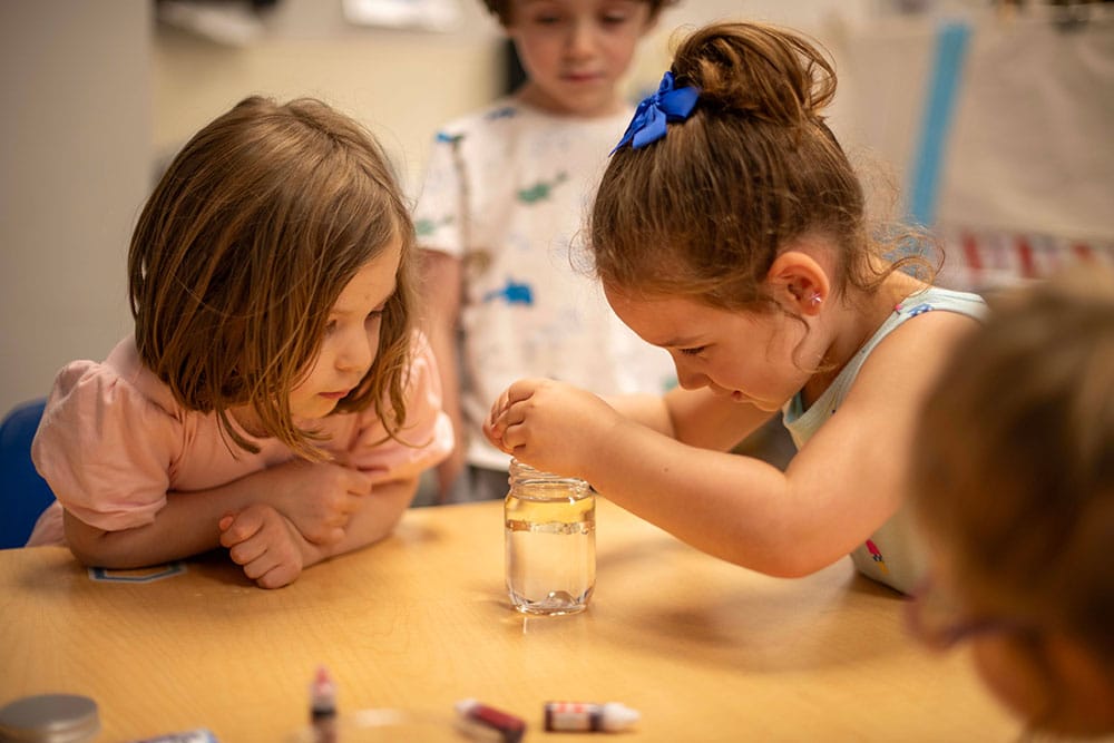 Three young children sit at a table. One girl is stirring a clear glass of water while another girl and a third child watch. Food coloring bottles and a small dropper are on the table.