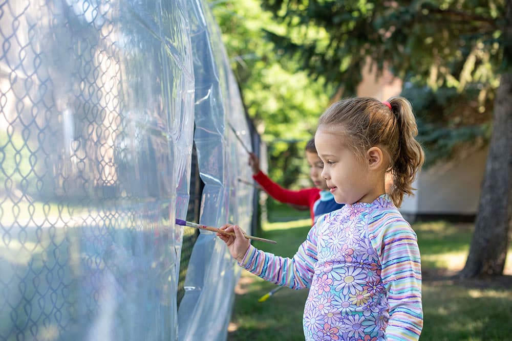 Two young children outdoors paint on a transparent plastic surface stretched across a fence. Trees and grass are visible in the background. One child in focus wears a flower-patterned shirt and holds a paintbrush.