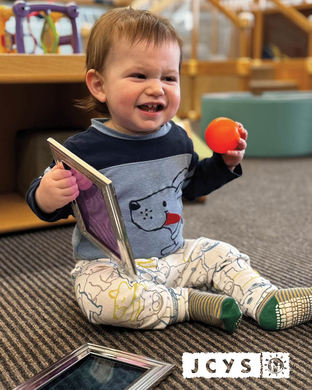 A smiling toddler sits on a carpeted floor holding a framed photo in one hand and an orange ball in the other. Shelves and toys are visible in the background. The image has a white JCYS logo at the bottom right.