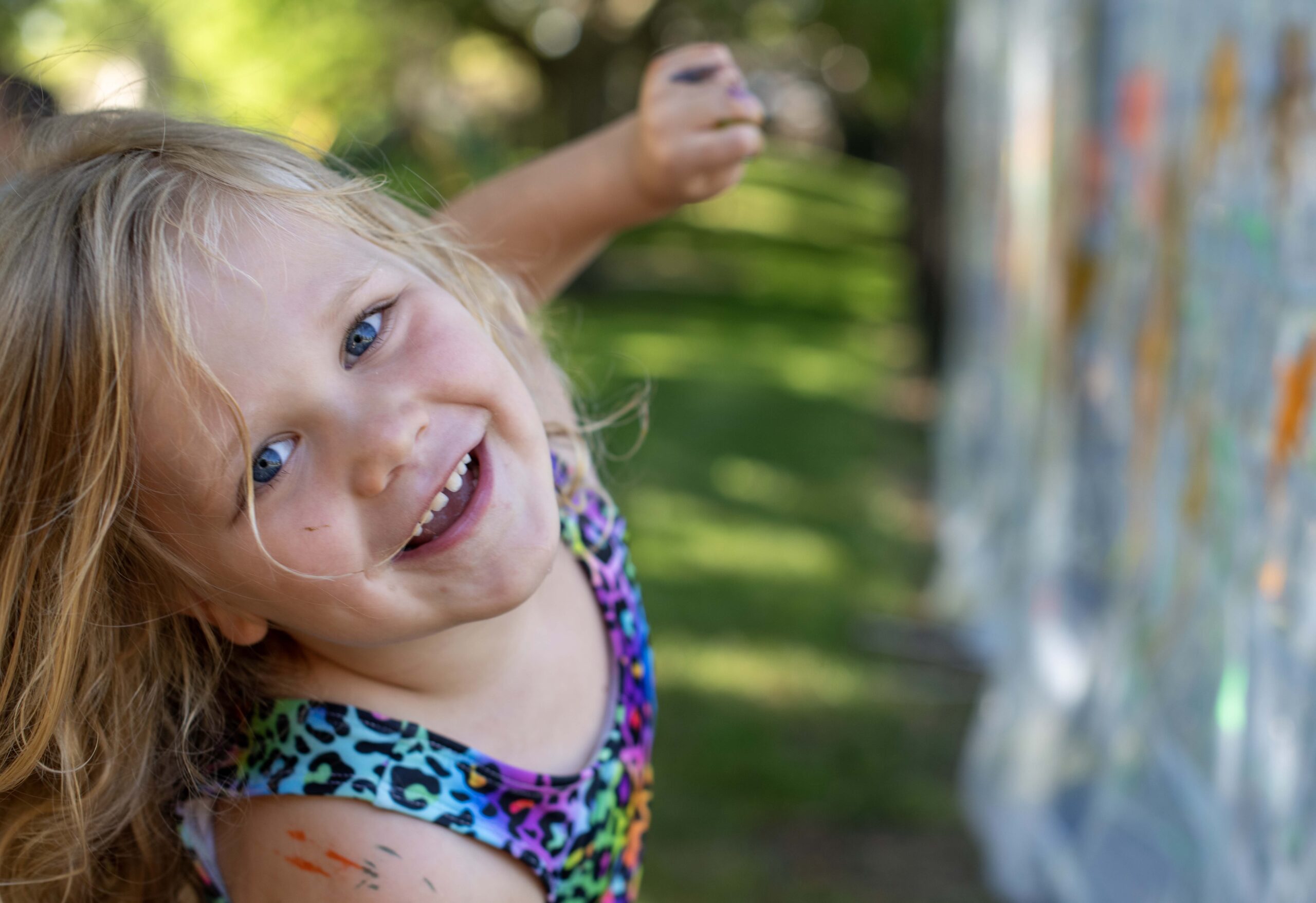 Young child with blonde hair smiles at the camera while painting on an outdoor surface. The child wears a colorful, leopard-print top, and there are paint marks visible on their arm and face. Green grass and trees are in the background.