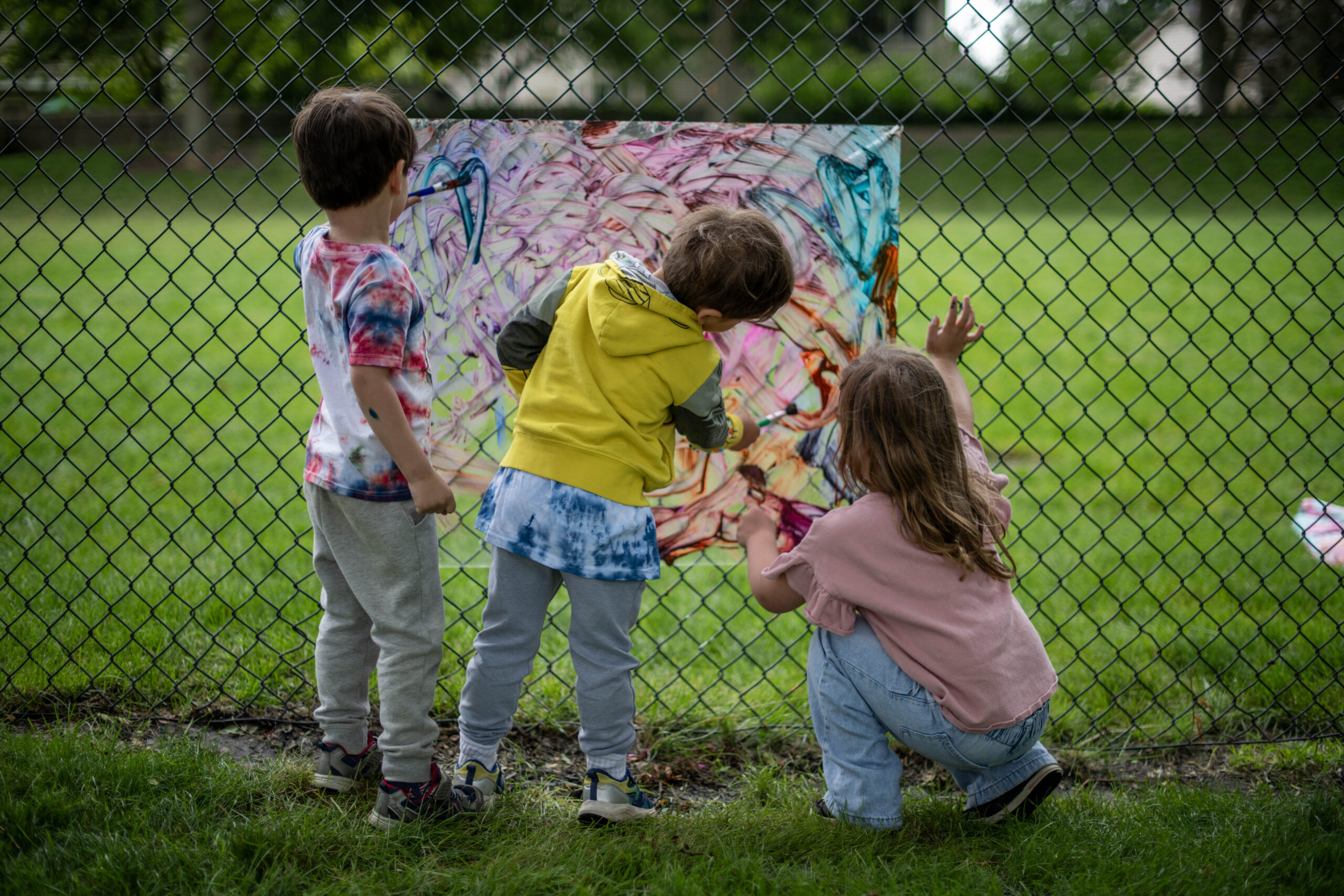 Two men stand under a canopy speaking to a large group of seated children outdoors. There is a fenced area, a shed, and some equipment in the background. The event appears to take place in a sunny courtyard.