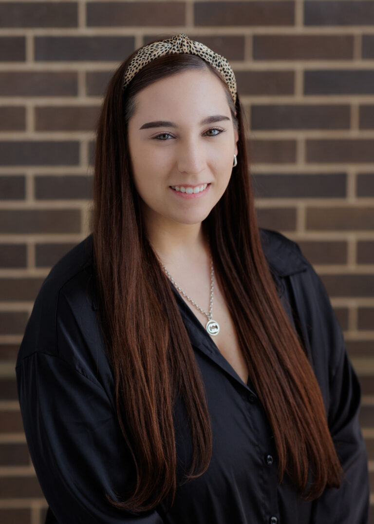 A woman with long brown hair wearing a leopard-print headband and a black collared shirt smiles at the camera in front of a brick wall.