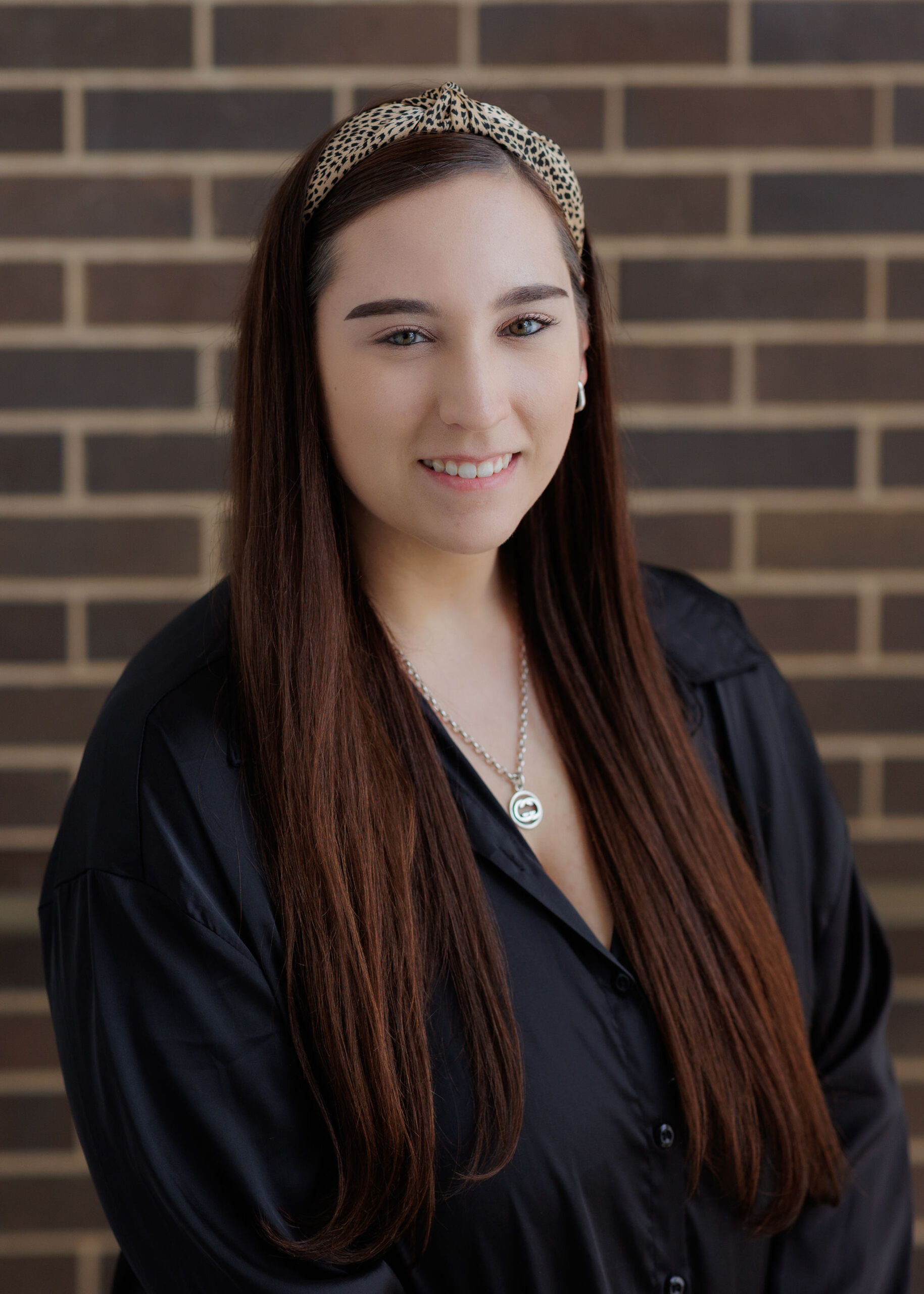 A woman with long brown hair wearing a leopard-print headband and a black collared shirt smiles at the camera in front of a brick wall.