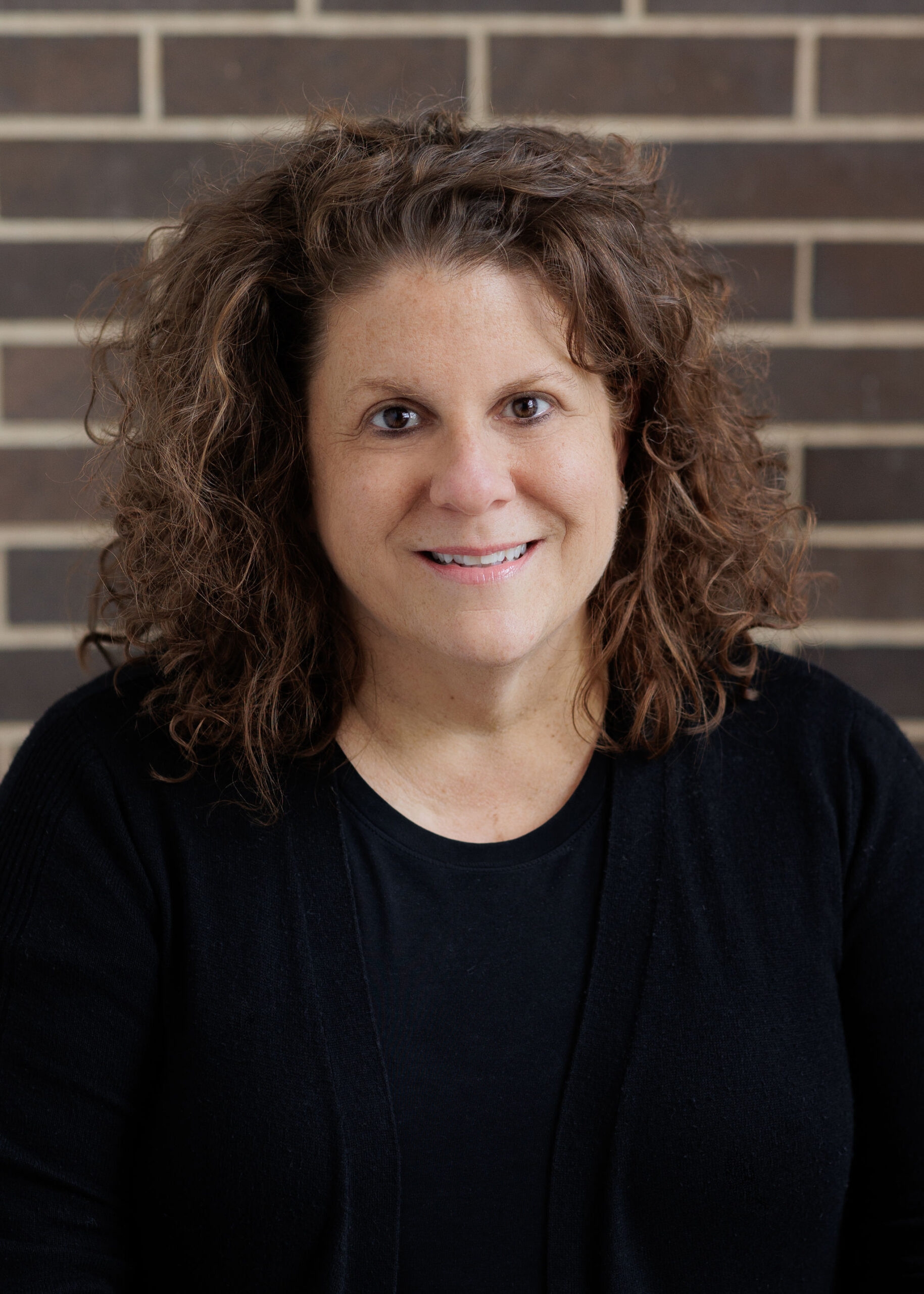 A woman with shoulder-length curly brown hair wearing a black shirt, posing in front of a brick wall.