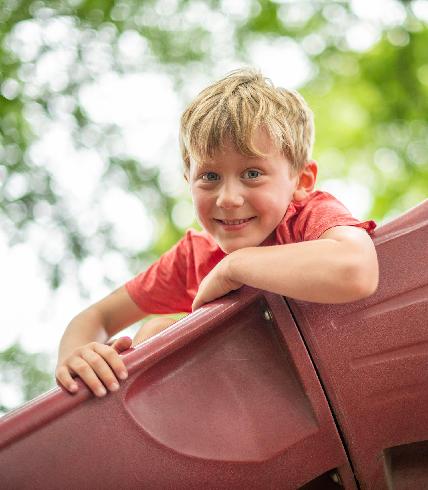 A young child in swimwear stands on grass, holding a colorful water blaster. A blue plastic kiddie pool with water is nearby. Raised garden beds and a chain-link fence are in the background. The scene is outdoors and sunny.