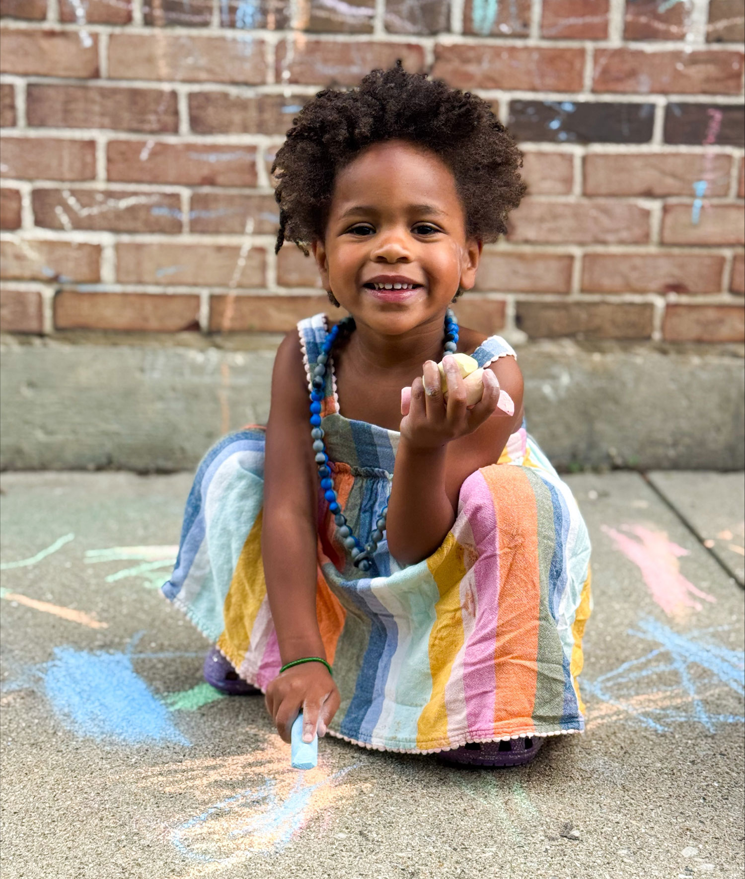 A young child in swimwear stands on grass, holding a colorful water blaster. A blue plastic kiddie pool with water is nearby. Raised garden beds and a chain-link fence are in the background. The scene is outdoors and sunny.