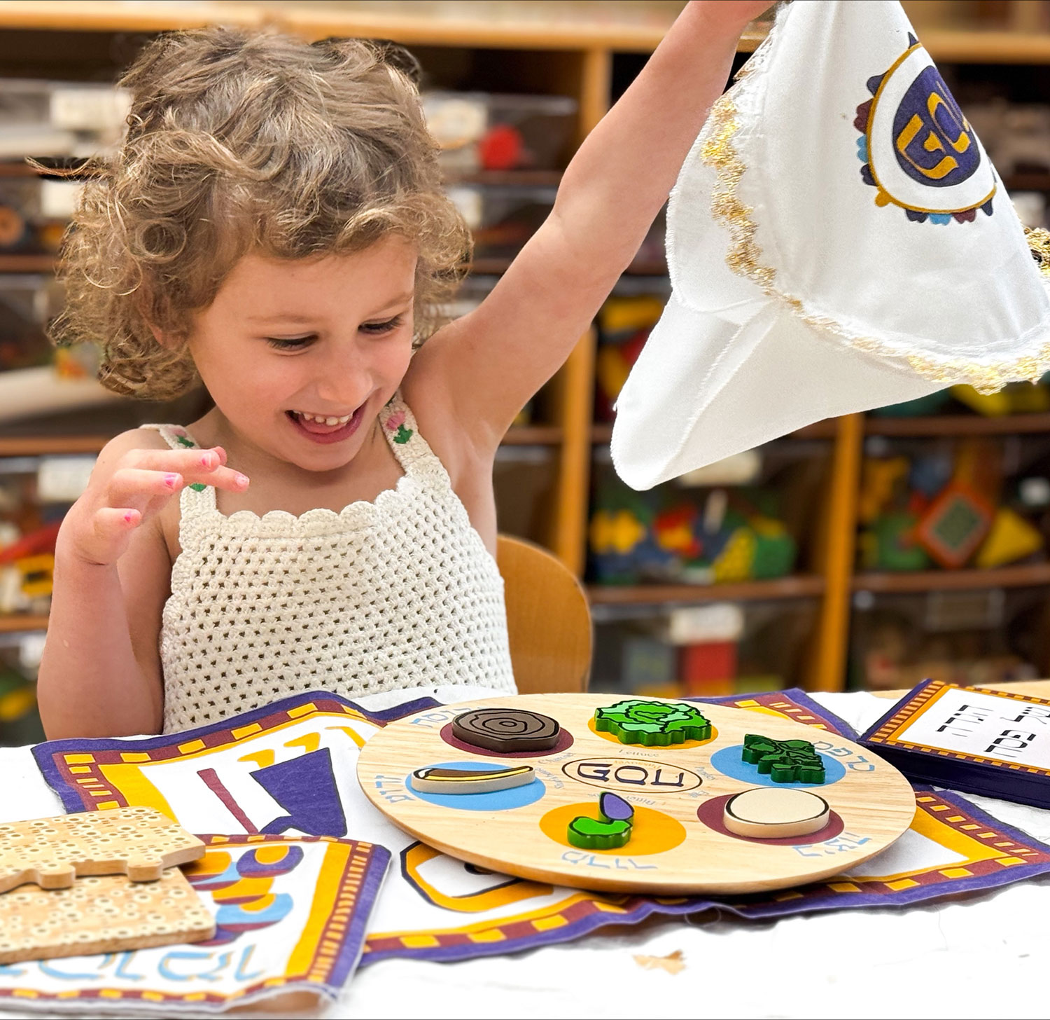A baby wearing a striped shirt crawls on a play mat indoors. A plush giraffe toy is on the mat beside the baby. Shelves and chairs are visible in the background.