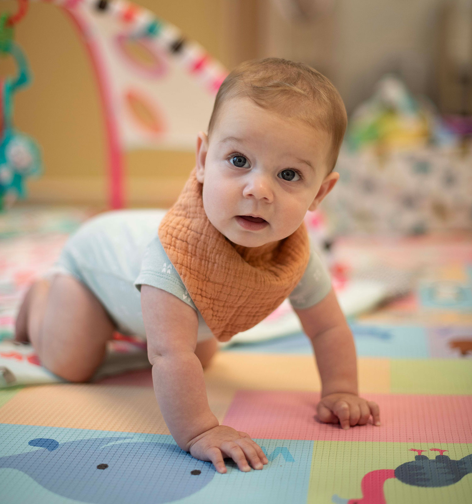 A baby wearing a striped shirt crawls on a play mat indoors. A plush giraffe toy is on the mat beside the baby. Shelves and chairs are visible in the background.