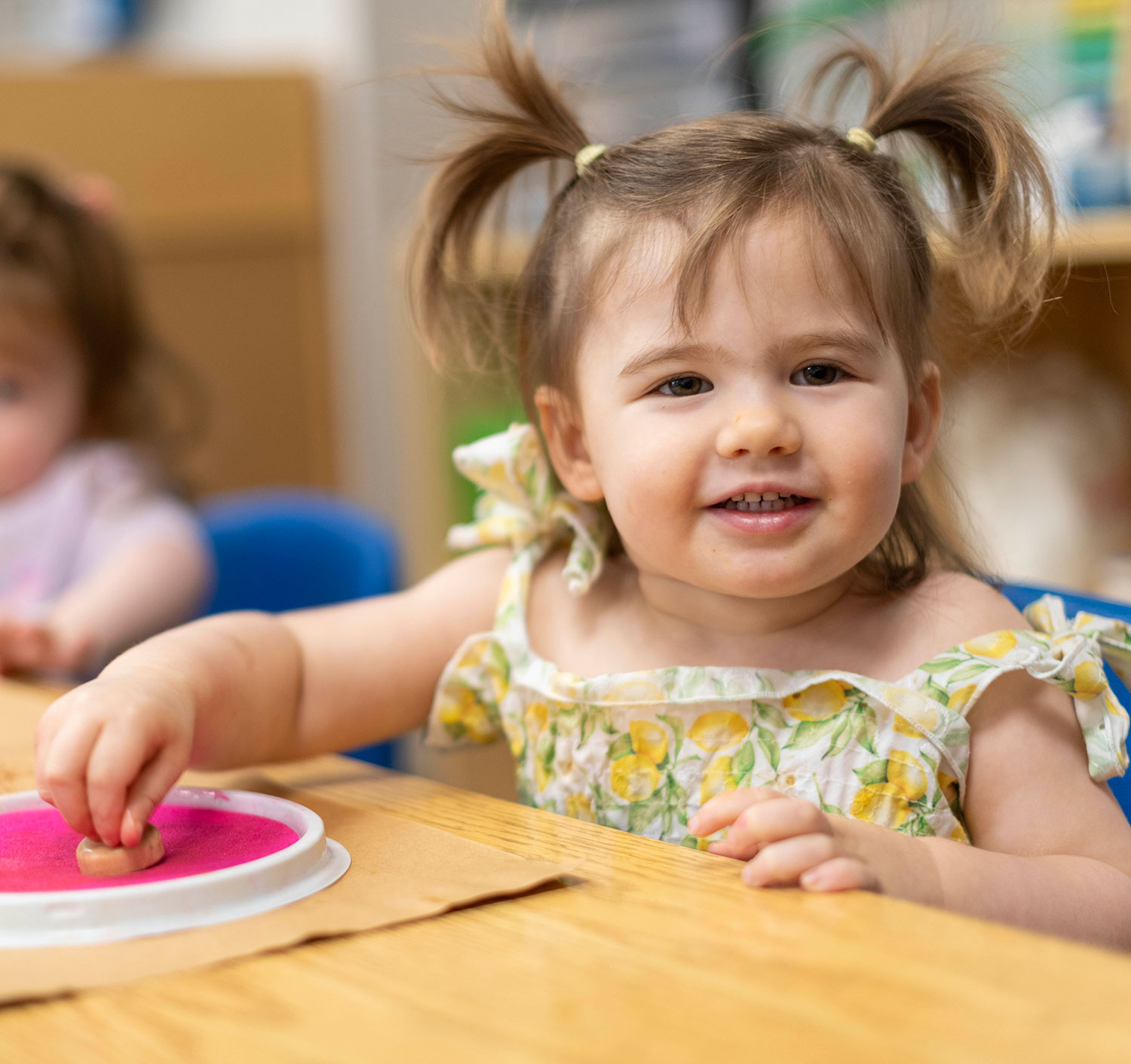 A baby wearing a striped shirt crawls on a play mat indoors. A plush giraffe toy is on the mat beside the baby. Shelves and chairs are visible in the background.