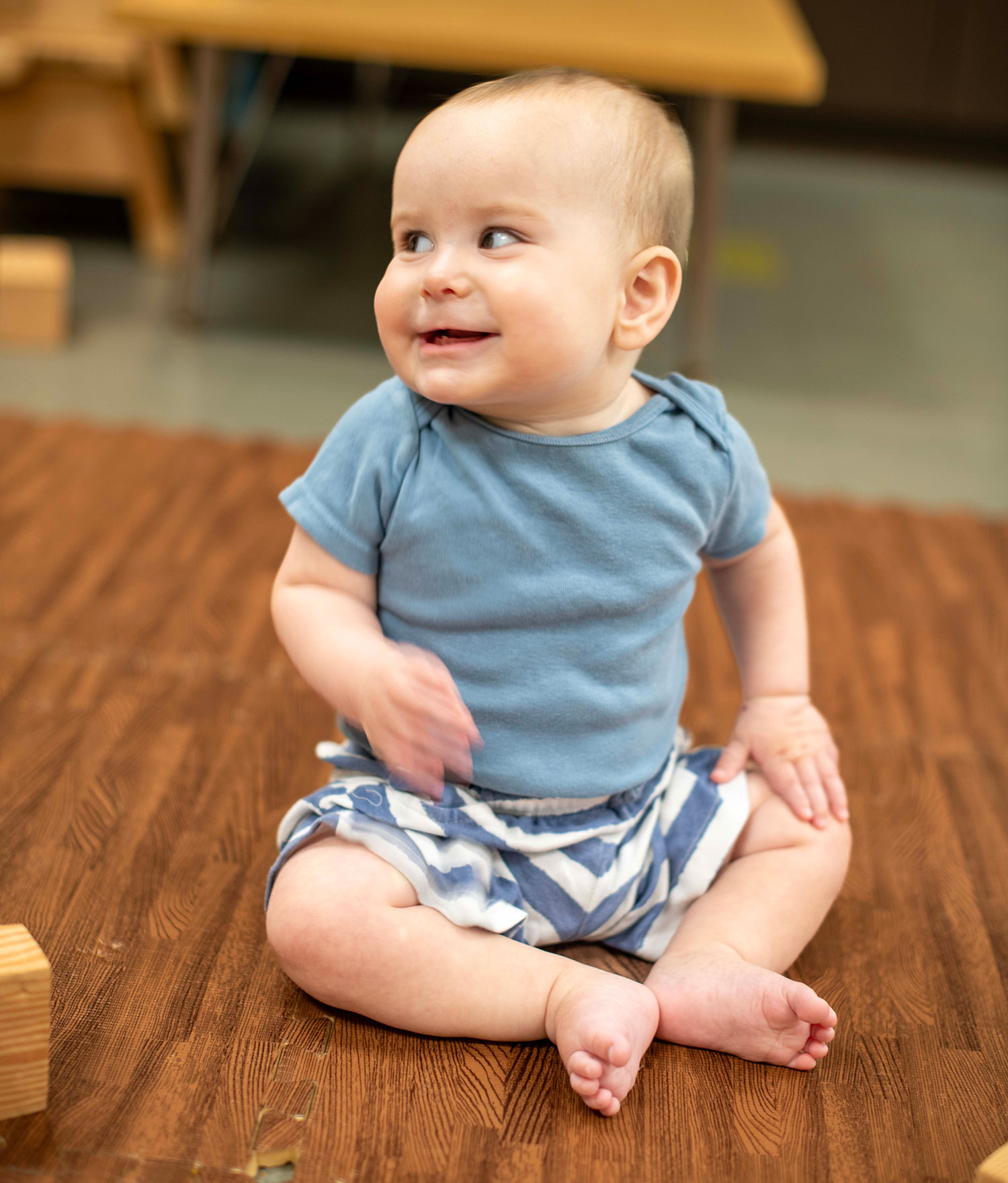 A baby wearing a striped shirt crawls on a play mat indoors. A plush giraffe toy is on the mat beside the baby. Shelves and chairs are visible in the background.