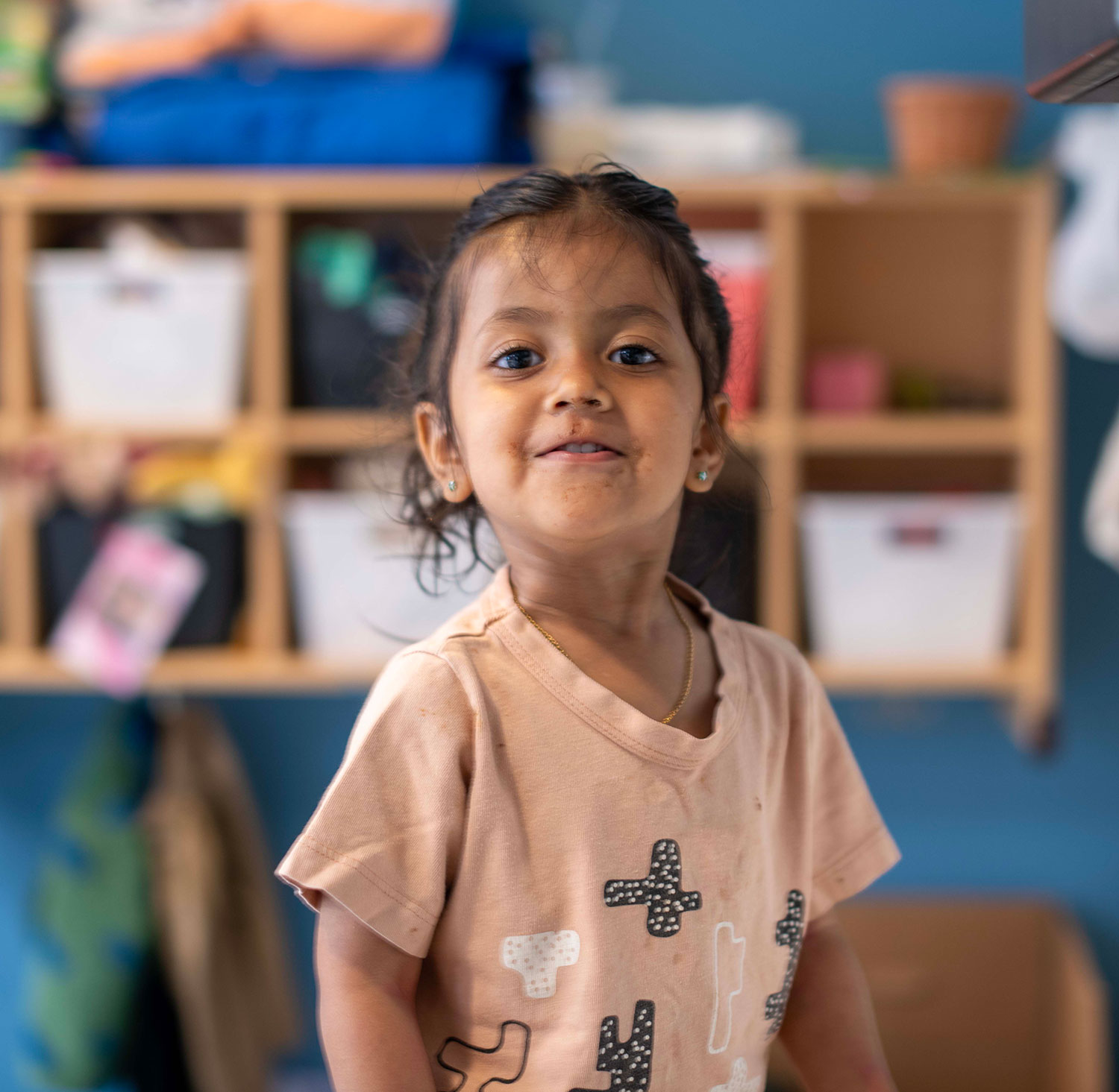 A baby wearing a striped shirt crawls on a play mat indoors. A plush giraffe toy is on the mat beside the baby. Shelves and chairs are visible in the background.