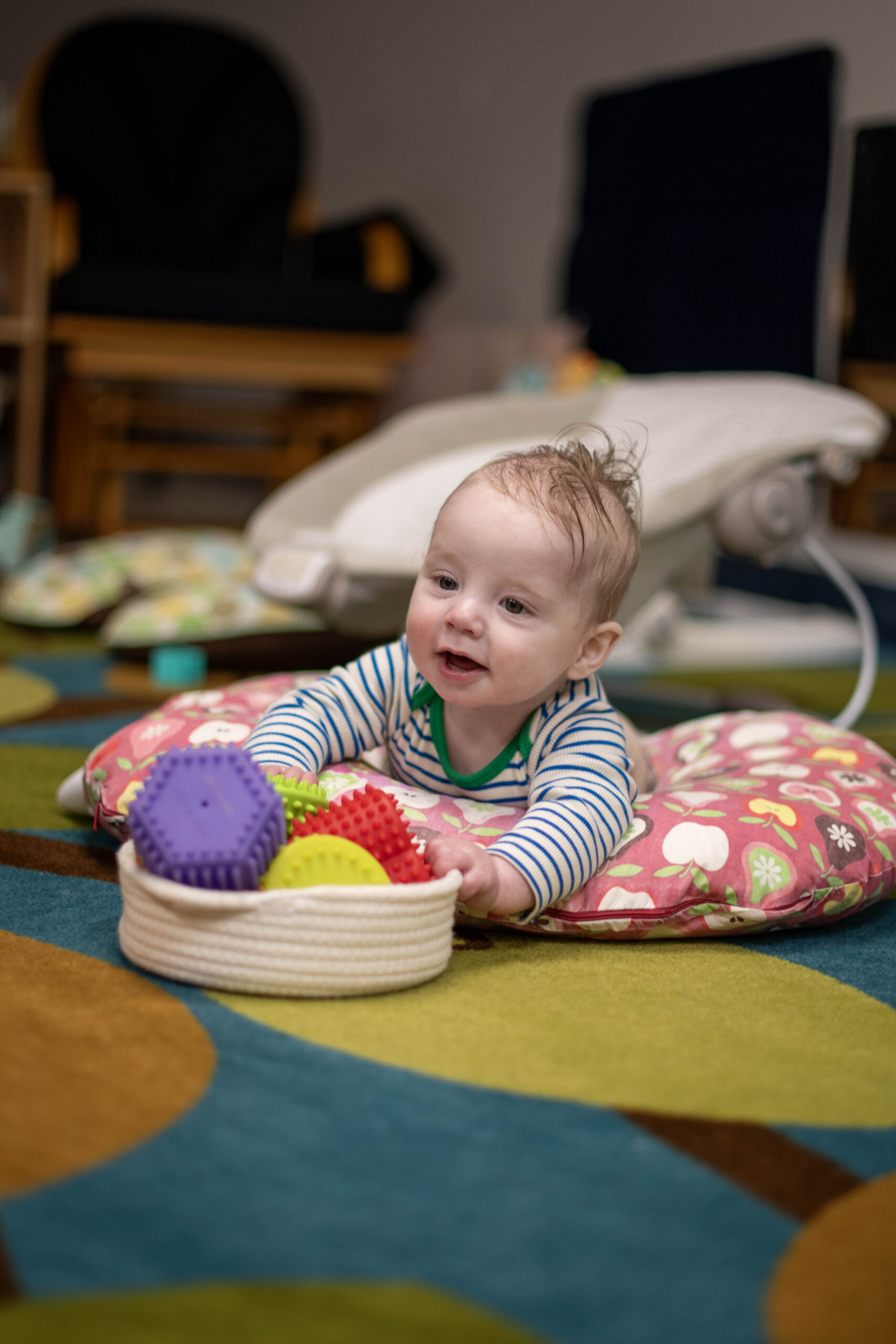 A baby wearing a striped shirt crawls on a play mat indoors. A plush giraffe toy is on the mat beside the baby. Shelves and chairs are visible in the background.