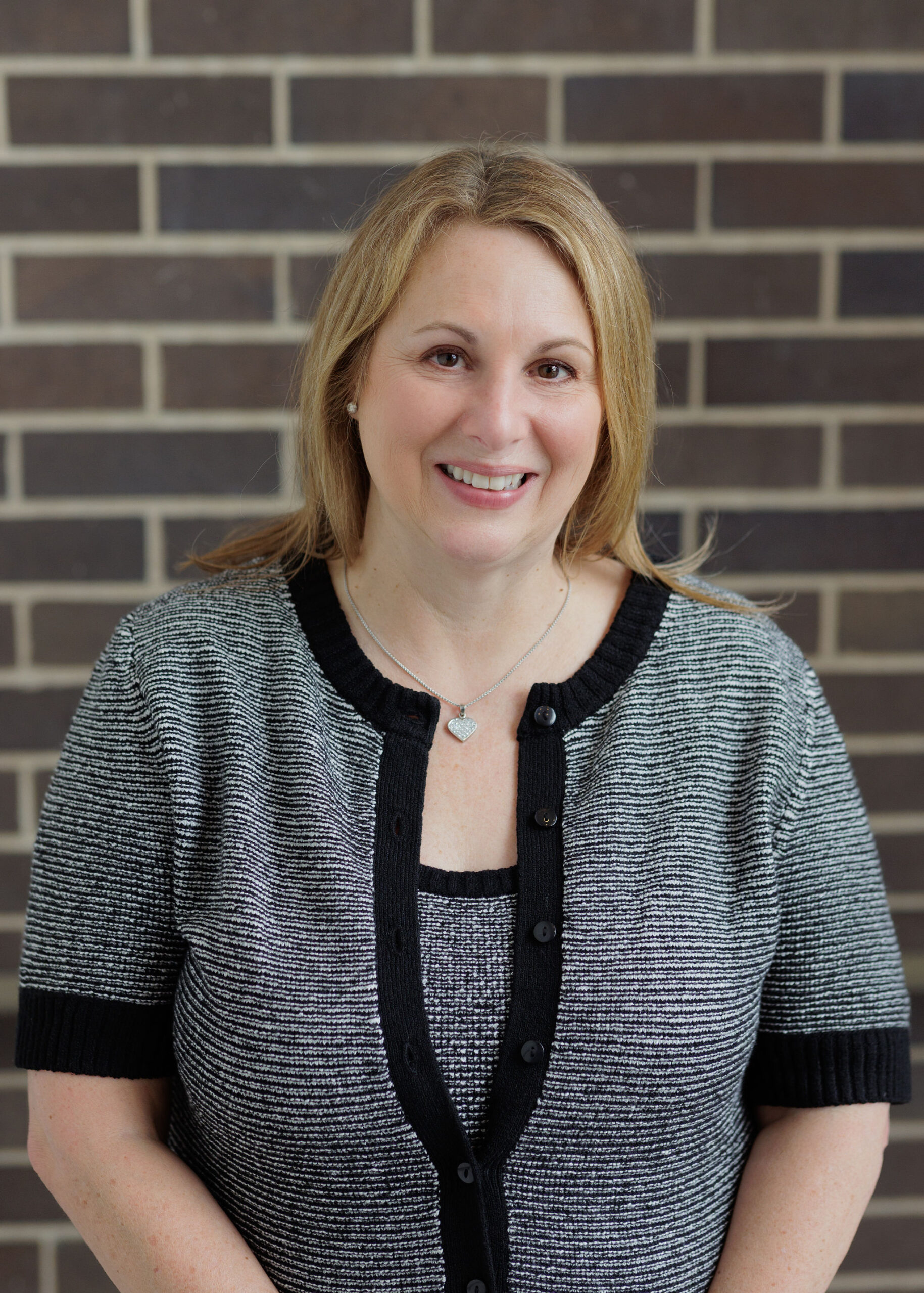 A smiling woman wearing a black and white patterned cardigan against a brick wall.