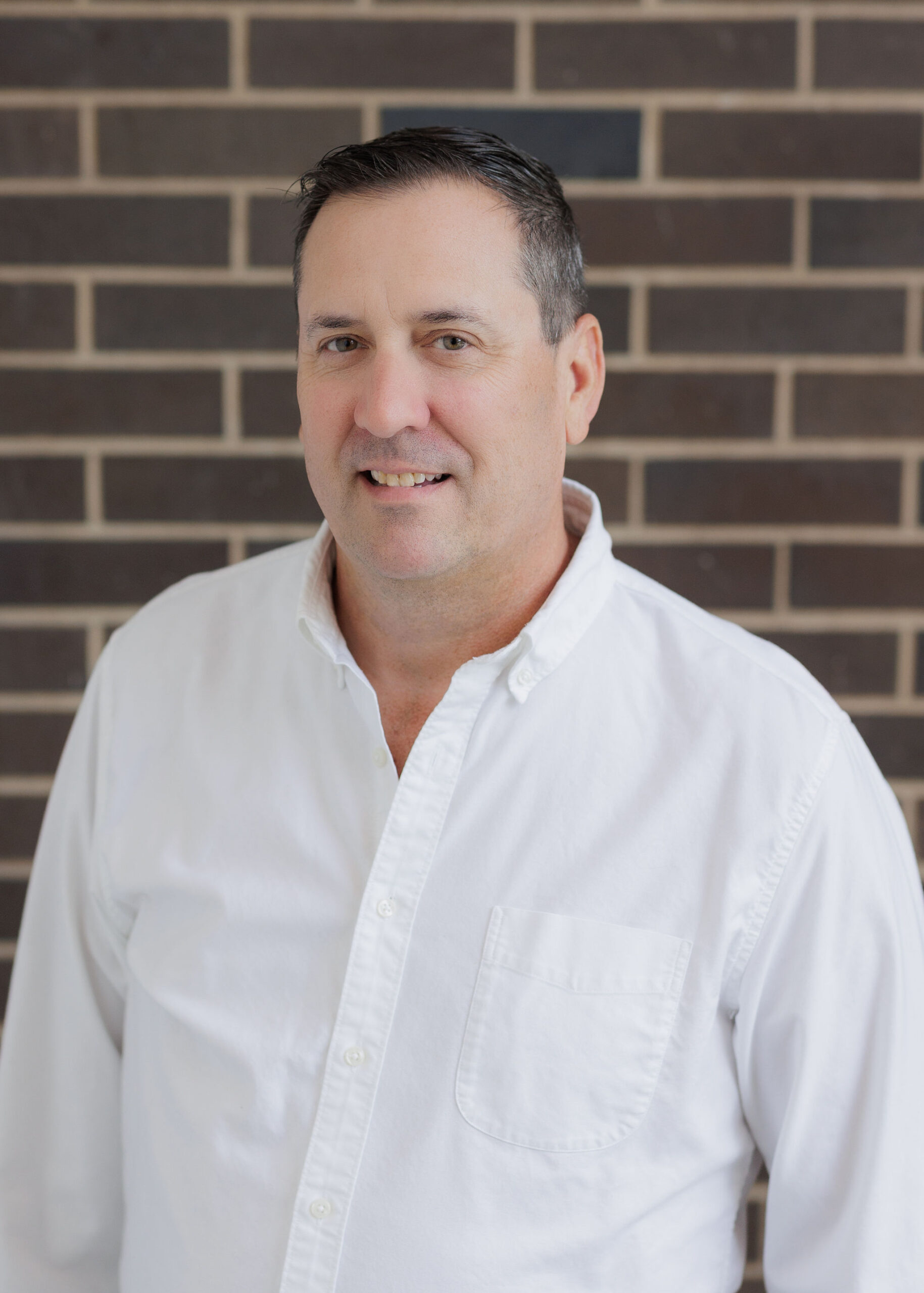 A man in a white button-down shirt stands in front of a brick wall.