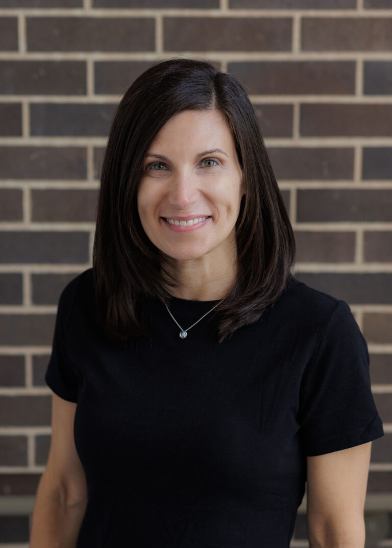A woman with shoulder-length dark hair smiles at the camera against a brick wall.