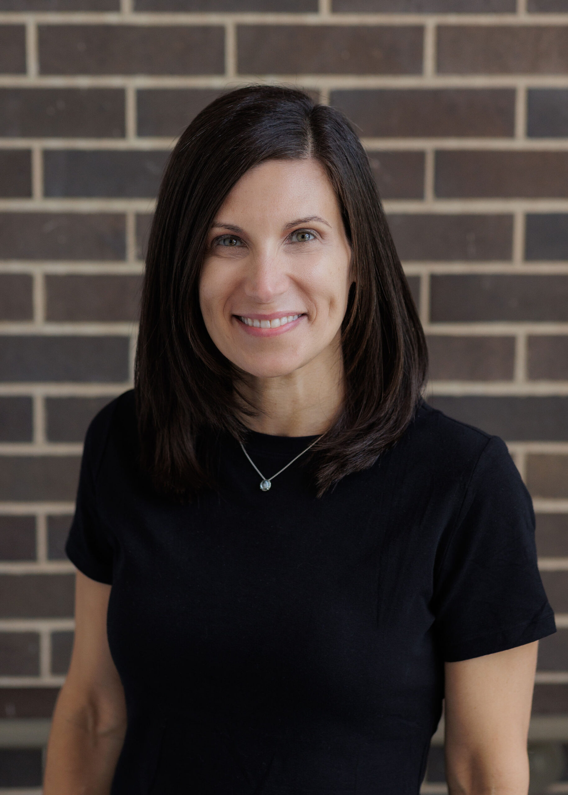 A woman with shoulder-length hair smiles in front of a brick wall, wearing a maroon top with button details on the shoulders.