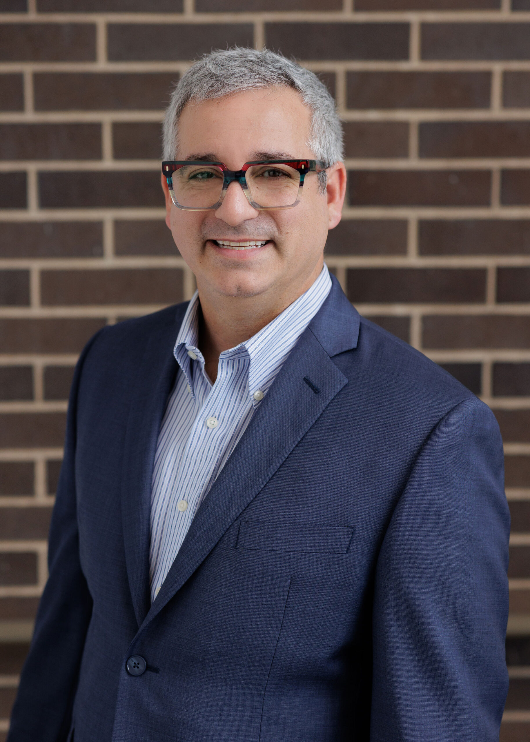 A man with graying hair and glasses wearing a blue suit jacket and striped button-down shirt stands in front of a brick wall.