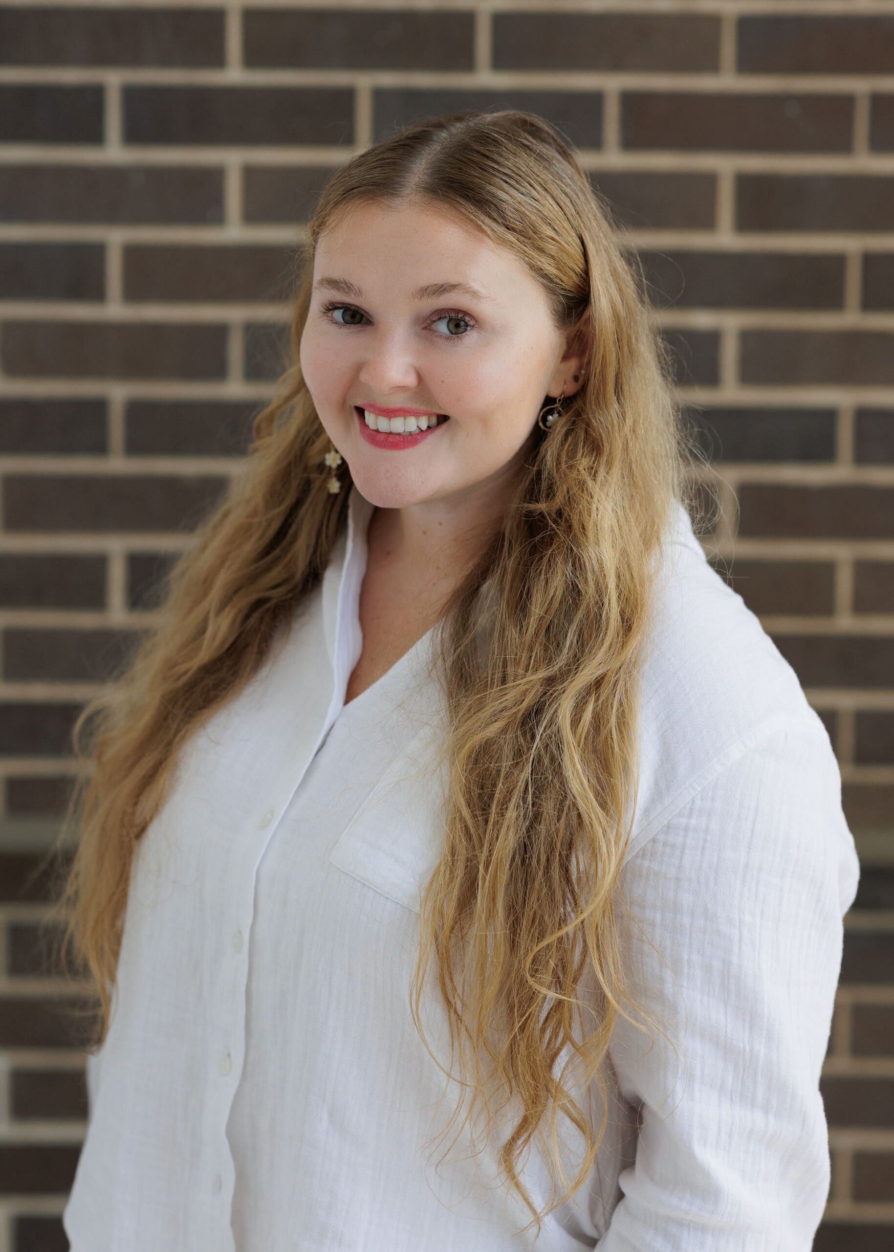 A woman with long blonde hair smiles while wearing a white collared shirt in front of a brick wall.