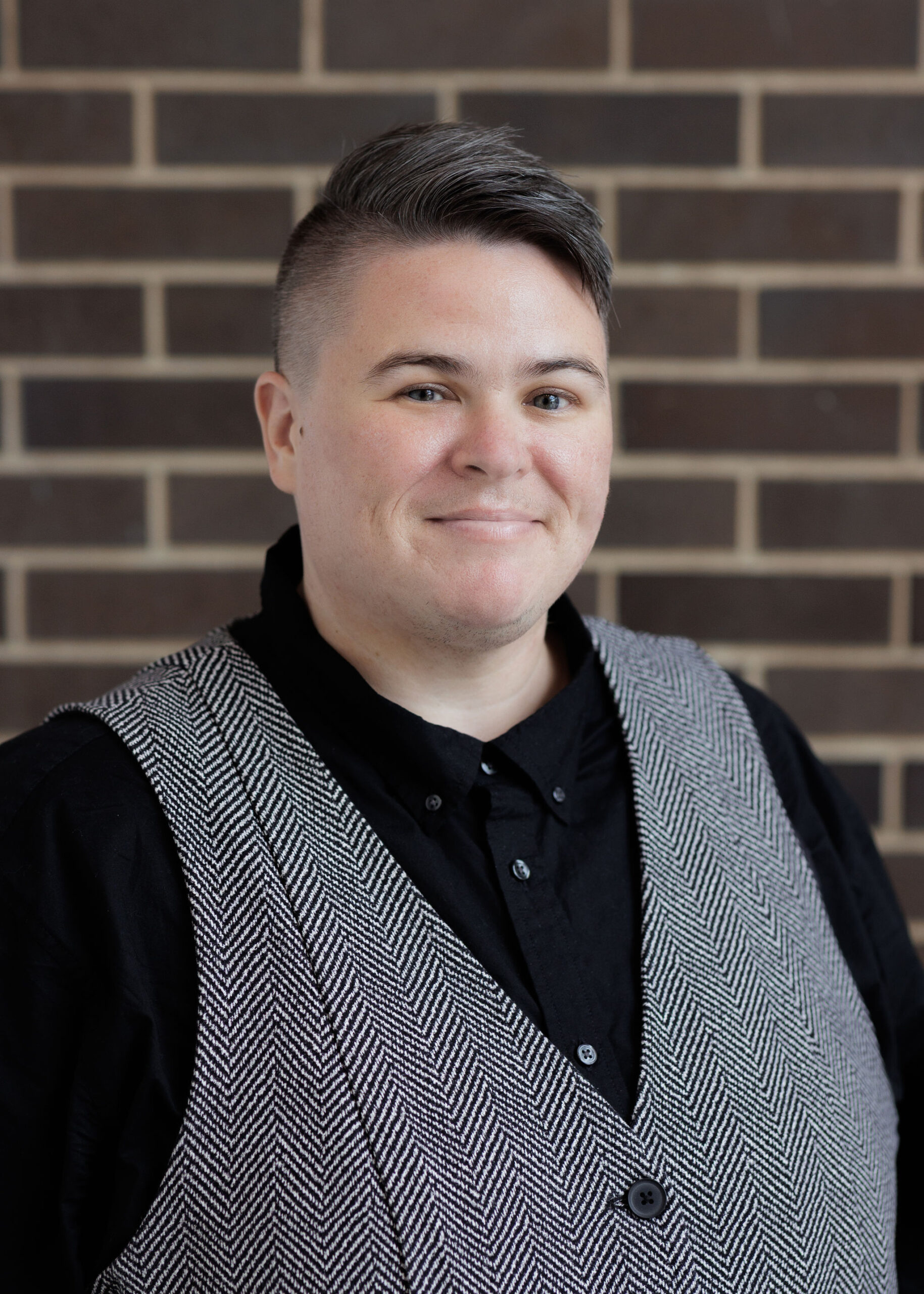 A person with short, faded hair wearing a black button-down shirt and a grey patterned vest stands in front of a brick wall.