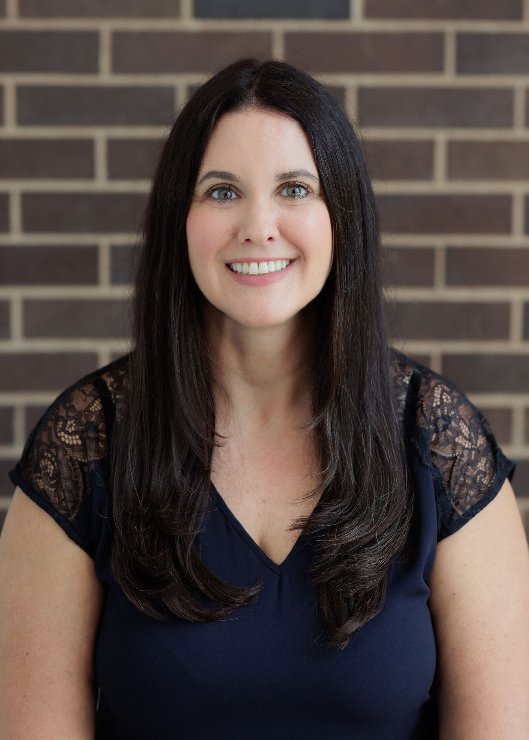 A smiling woman with long dark hair wearing a dark blue top against a brick wall.