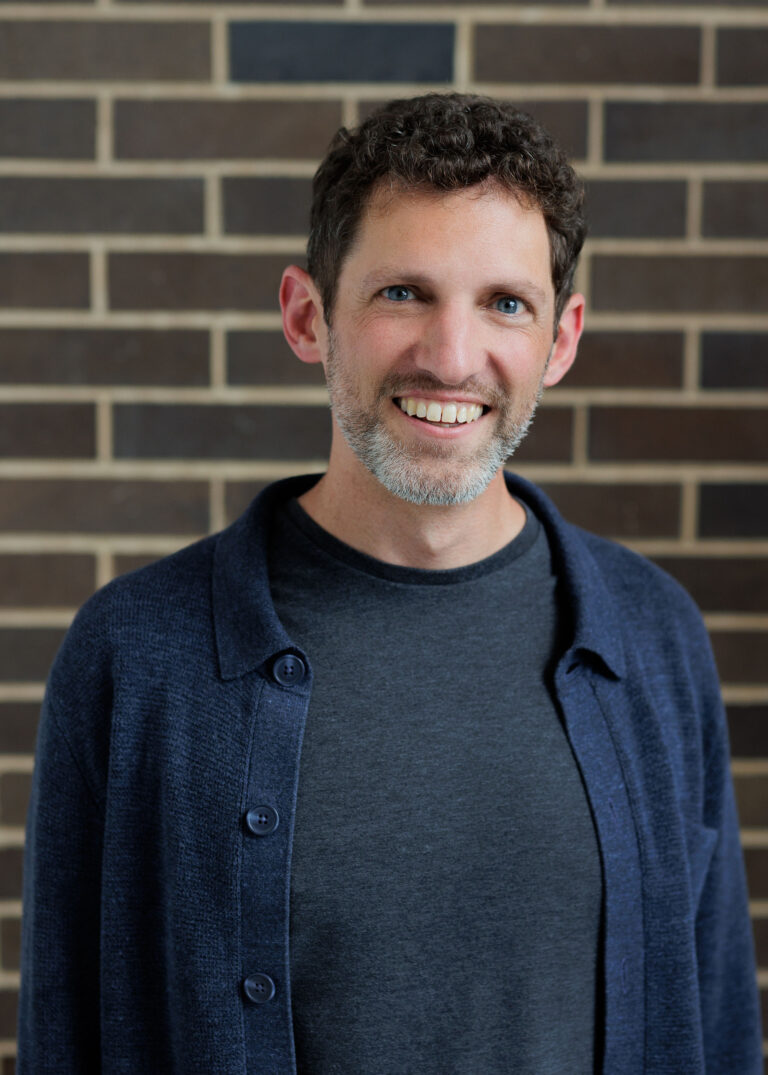 A smiling man with a short beard wearing a dark gray t-shirt and a blue cardigan against a brick wall.