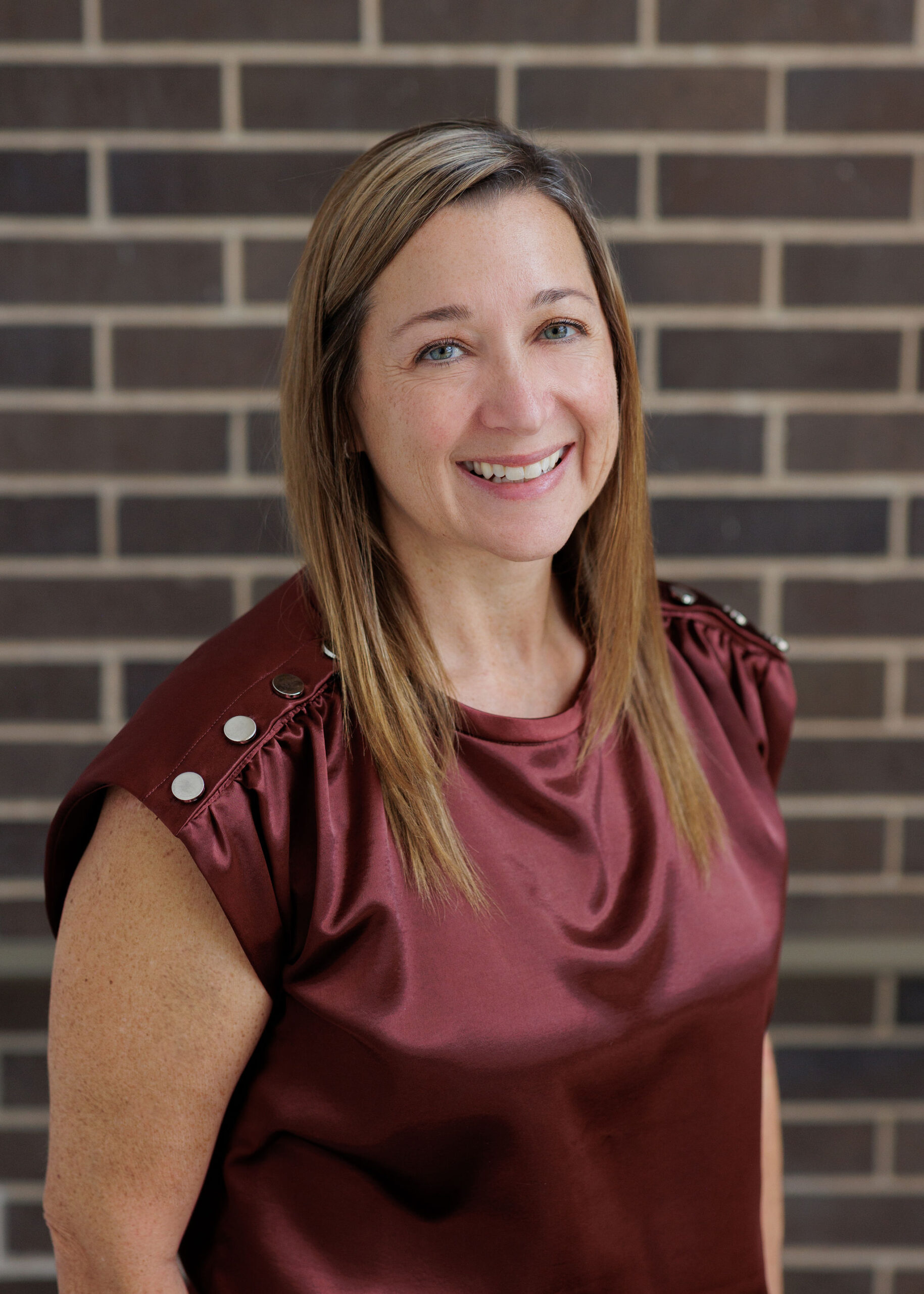 A woman with shoulder-length hair smiles in front of a brick wall, wearing a maroon top with button details on the shoulders.