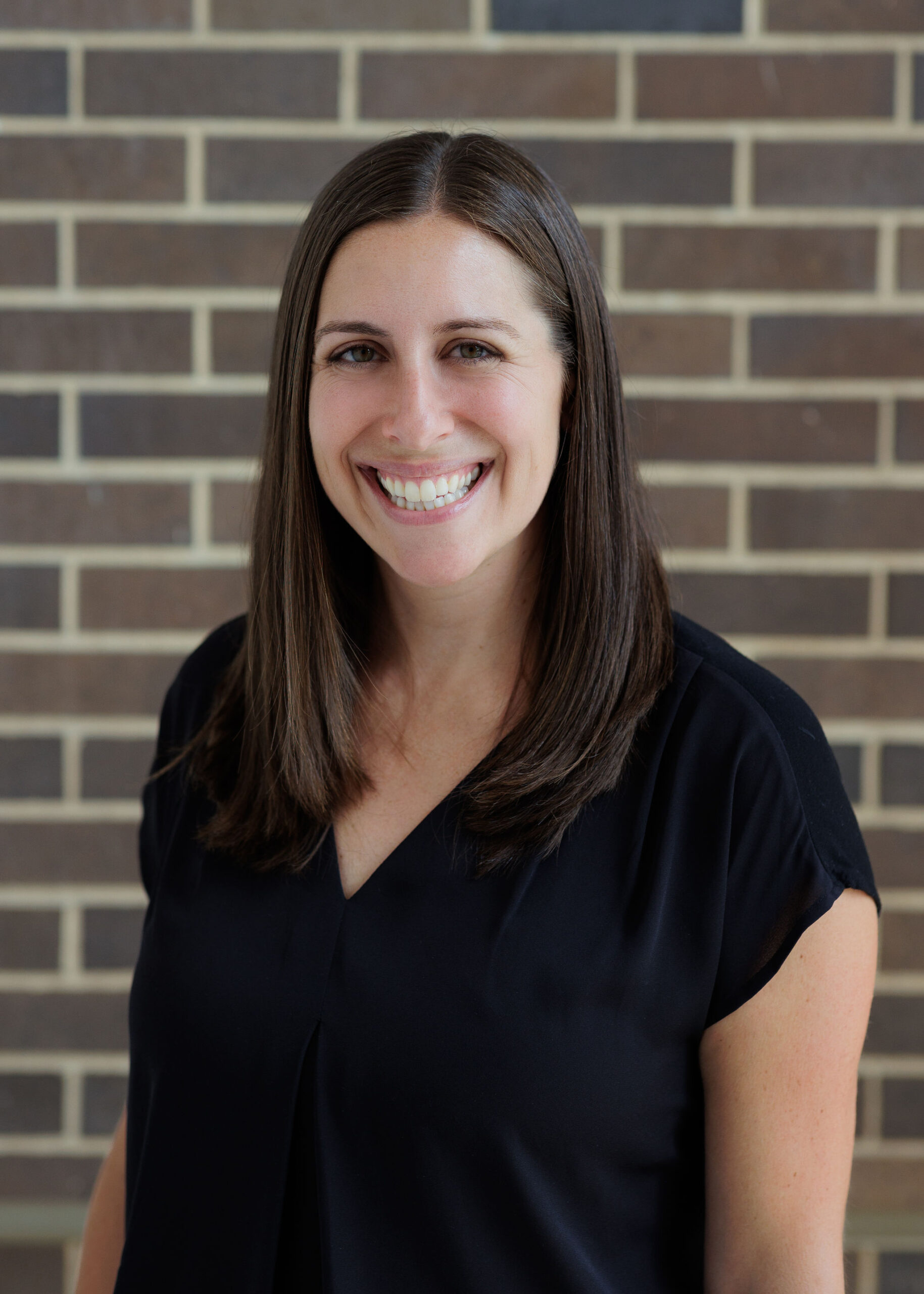 A smiling woman with long brown hair wearing a black top stands in front of a brick wall.