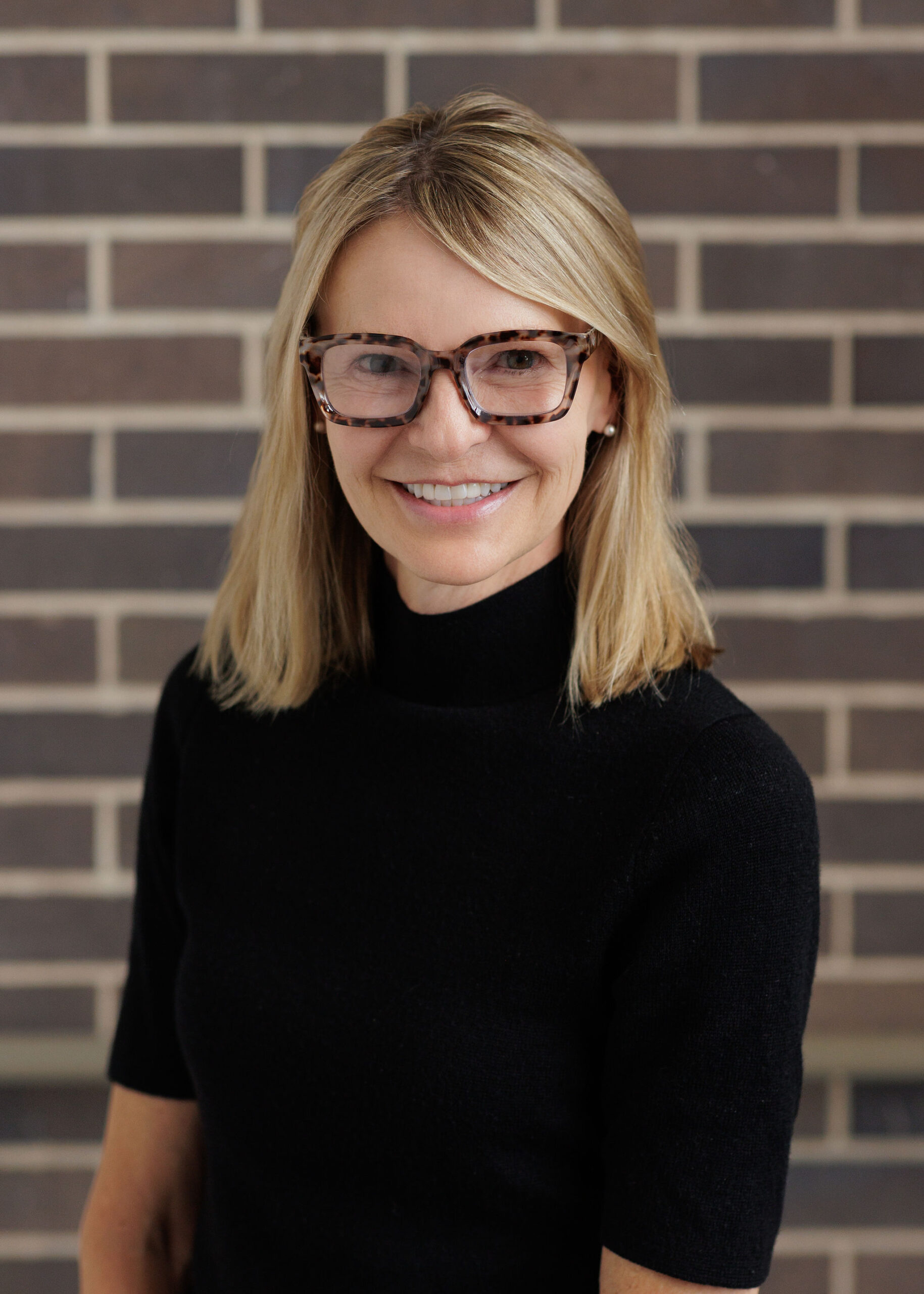A blonde woman wearing a black turtleneck and tortoiseshell glasses smiles at the camera in front of a brick wall.