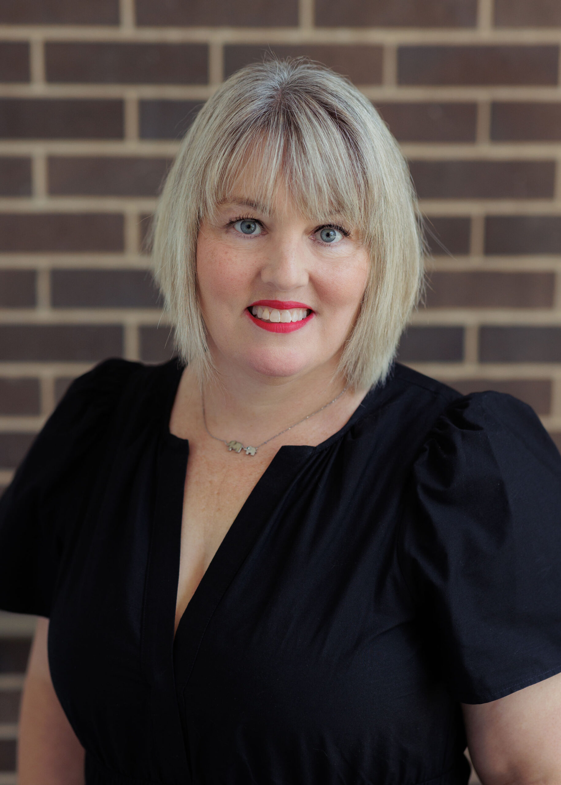 A woman with shoulder-length blonde hair wearing a black blouse and a small elephant necklace poses in front of a brick wall.