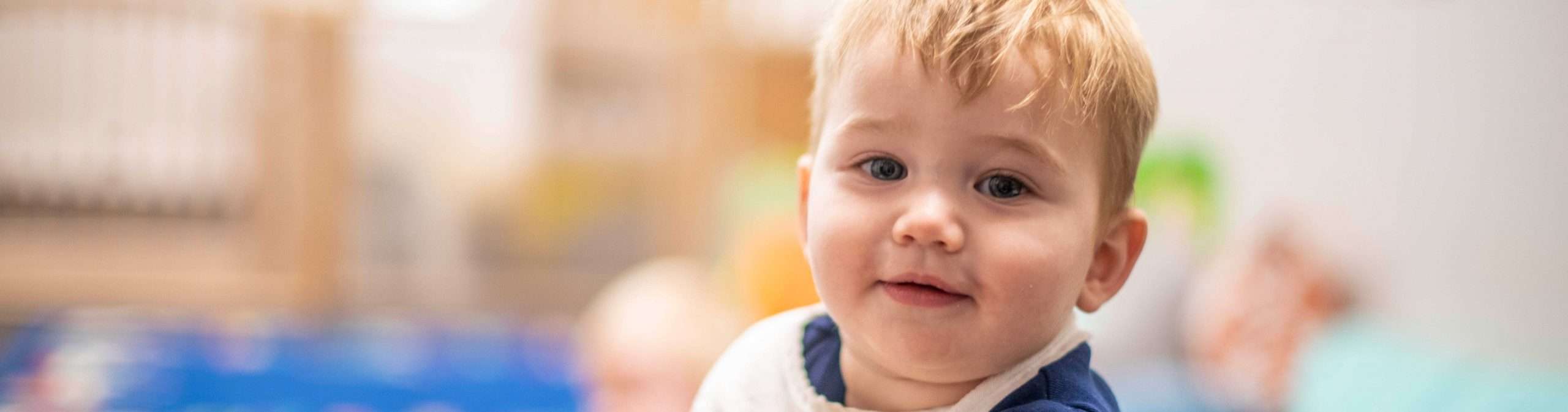 Blonde infant is smiling in classroom. He has a white bib and navy t-shirt on.