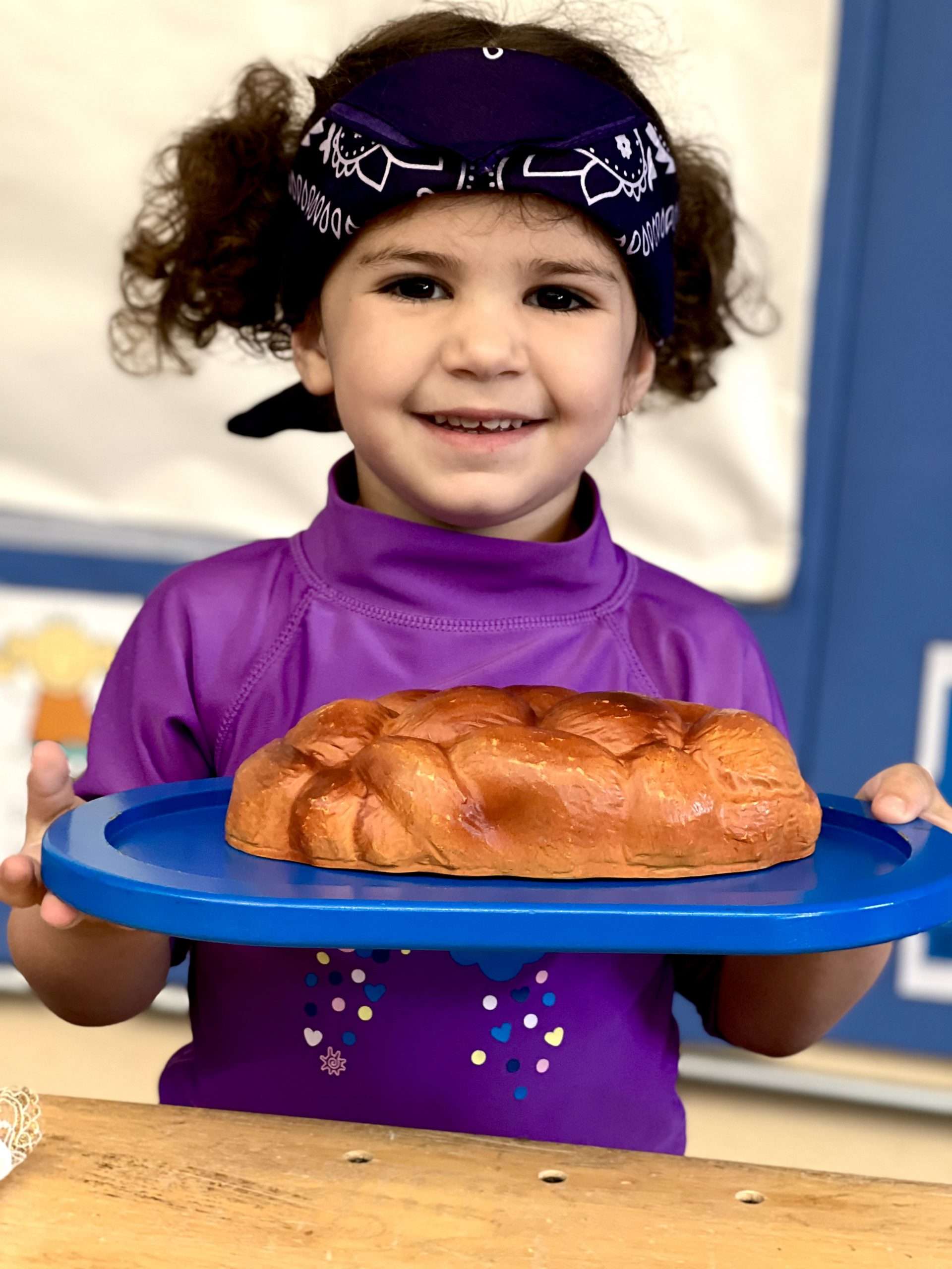 Child smiling and holding a freshly baked challah on a tray.