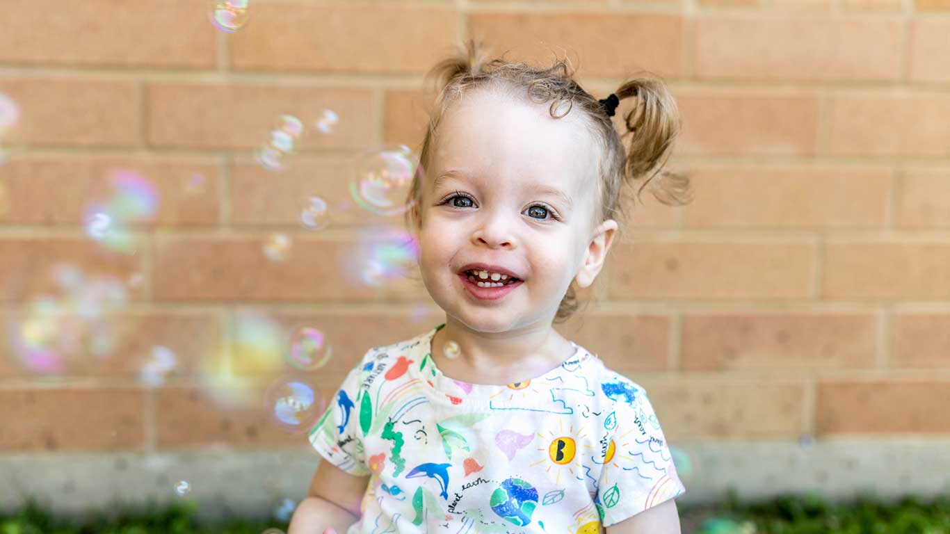 Child outside smiling at camera with bubbles floating on the left side.